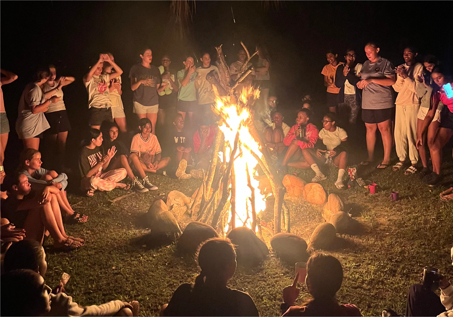 Large group of campers gathered around a glowing campfire at night, sharing stories and songs at Camp Anerley.