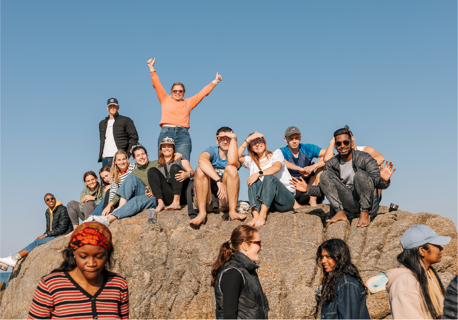 Camp Anerley group smiling and relaxing on a large rock under a clear blue sky during a beach outing.