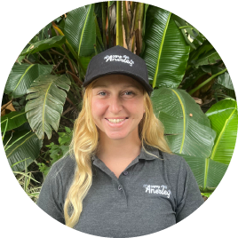 Smiling Camp Anerley team member wearing a grey Camp Anerley shirt and black cap, standing in front of large green tropical leaves.