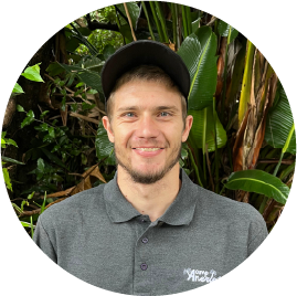 Smiling Camp Anerley team member wearing a grey Camp Anerley shirt and black cap, standing in front of green foliage.