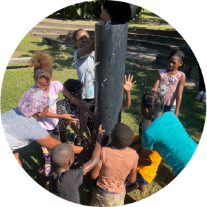 Children working together on a water activity at Camp Anerley, pouring water into a large pipe during an outdoor team challenge.